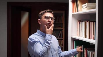 Thoughtful male student choosing a book from a white bookshelf in a library, stroking his chin while making a decision about his next reading video