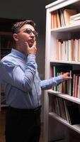 vertical A man in glasses is standing in front of a bookcase choosing a book video