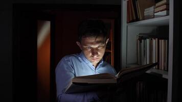 Focused man reading a large book illuminated by its pages in a dark room next to a white bookshelf at night, enjoying the silence and knowledge video