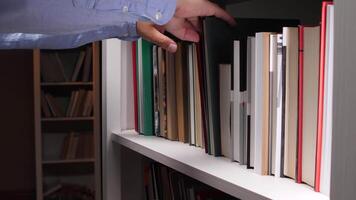 Close-up of a librarian's hands carefully placing a dark green hardcover book onto a white bookshelf filled with various colorful books, creating a vibrant and organized display of literature video