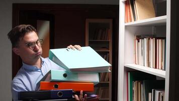 Young office worker putting a folder on a shelf in the office, he is holding other folders in his other hand, he is wearing glasses and a blue shirt video