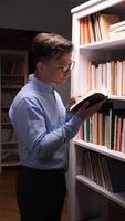 vertical A man in glasses is standing in front of a bookcase reading book video