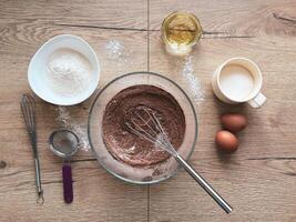 Glass bowl with flour and cocoa on wooden table with chocolate dough ingredients arranged around. photo