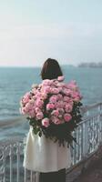 Woman Holding a Large Bouquet of Pink Roses While Standing by the Sea During a Calm Day photo