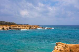 Coastal view of turquoise waters meeting golden cliffs with small caves, rocky formations, and a tree lined horizon under a partly cloudy sky. photo