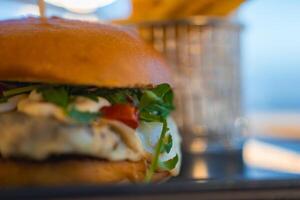 A golden bun burger with arugula, cheese, and tomato, paired with fries in a metal basket. Soft lighting and a hint of a coastal view in Cyprus. photo