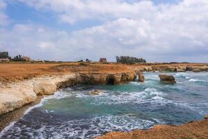 White cliffs with natural sea carved caves meet turquoise waters along a rocky shoreline in Cyprus, with scattered buildings and trees in the background. photo