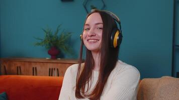 Overjoyed young woman in wireless headphones dancing, singing on couch in living room, choosing favorite energetic disco music in smartphone application, entertaining indoors. People relax activities video