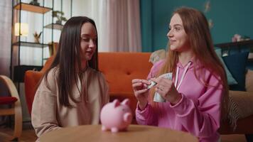 Happy girls friends siblings sitting on floor and take turns dropping dollar banknote into piggy bank, save pocket cash for fun, dreams, future vacation. Keep money, thinking about tomorrow concept video