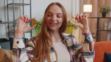 Woman freelancer holding two gold coins at home, successful developer programmer. Girl stock trader earning bitcoins after online monitoring trading operations. Increasing wealth, financial prosperity video