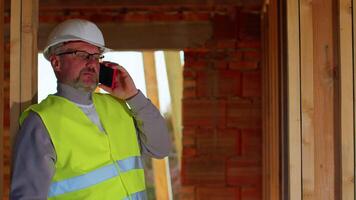 Civil engineer architect specialist talking on mobile phone to control working at construction site. House on background. Developer builder in helmet inspecting building. Real estate home project video