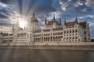 The neo Gothic Hungarian Parliament Building stands by the Danube River in Budapest, with sunlight streaming through clouds and calm waters in the foreground. photo