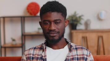 Lovely playful happy man african american smiling blinking eye, looking at camera with toothy smile, winking and flirting, expressing optimism. Guy sitting at home in living room on sofa, indoors video