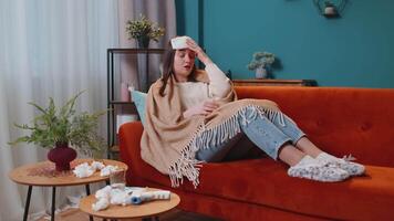 Portrait of young woman suffering from cold or allergy in plaid lying on sofa in living room at home. Sick adult girl with towel on head blows nose snot into napkin. Coronavirus quarantine, pandemic video