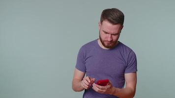 Handsome bearded adult man in purple t-shirt using mobile phone typing new post on web, sms message, browsing, addiction of social networks. Young one guy studio shot isolated on gray wall background video