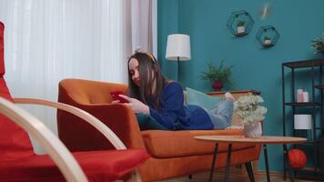 Relaxed smiling Caucasian young woman lying on sofa with smartphone in hands in living room home. Female girl tapping, scrolling, browsing online. Cheerful lady watching something on mobile phone video