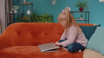 Joyful school kid girl use laptop computer sitting on sofa alone at home. Child learning reading online social media content, play education lessons game, chatting with friend. Children tech addiction video