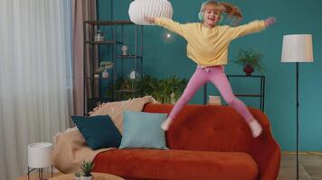Portrait of one Caucasian funny little happy child girl dancing on sofa while listening music in headphones in cozy living room. Small cute teen school kid at home alone jumping and moving to rythm video