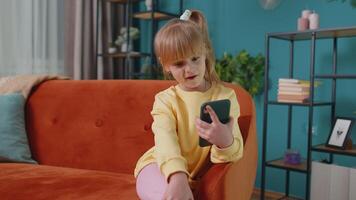 Portrait of Caucasian little happy child girl enjoying conversation on mobile phone with friend relaxing sitting on sofa in living room. School toddler kid at home alone. Communication teens concept video