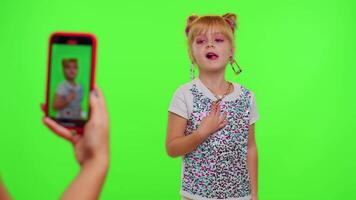 Lovely little Caucasian kid girl blogger front of phone camera record enjoy dance content on green chroma key background. Social distance coronavirus pandemic concept. Freedom active lifestyle video