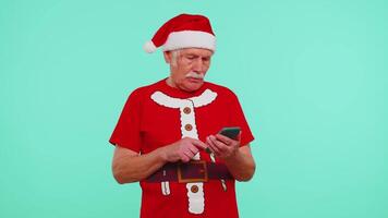 Grandfather in Christmas t-shirt using mobile phone typing new post on web, sms message, browsing, addiction of social networks. Blue background. Happy New Year celebration holidays shopping online video