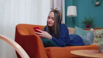 Young caucasian woman using smartphone while lying on couch at home. Smiling girl using spending time with cell gadget technology, relaxing, chatting, online shopping, watching on mobile phone video