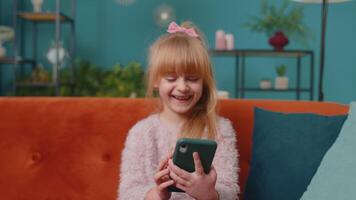 Happy little school kid girl using mobile phone technology device sitting on sofa alone at home. Child toddler hold smartphone watching funny cartoons, chatting with friends. Children tech addiction video