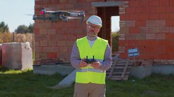 Male investor drone operator checks the work, controls an aircraft, holding remote controller. Professional builder in helmet and vest standing in front of construction site. House building. video