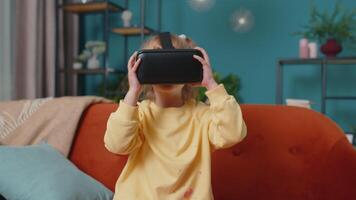 Exited amazed toddler girl sitting on sofa in living room using virtual reality futuristic technology VR app headset helmet to play simulation 3D 360 game, drawing. Teen school kid at home alone video