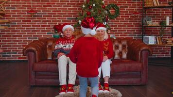 Girl set timer on mobile phone for taking multigenerational family selfie portrait while celebrating Xmas. Sending positive emotions sitting on sofa near Christmas tree at home room. Online call video
