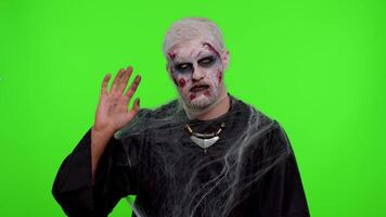 Creepy scary man with bloody scars face, Halloween zombie make-up. Scary wounded undead guy waves hand palm in hello gesture welcomes someone with hospitable expression expresses positive emotions video
