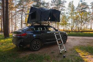 A modern SUV with a foldable rooftop tent set up in a forest clearing. A ladder extends to the ground, surrounded by tall trees and sunlight. photo