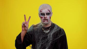 Frightening man with Halloween zombie bloody wounded makeup showing victory sign, hoping for success and win, doing peace gesture, smiling with kind optimistic expression on studio yellow background video