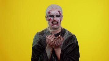 Upset disappointed zombie man with makeup with wounds scars wipes tears and cries from despair, being sad because of unfair things in yellow studio room. Sinister dead guy. Halloween, filming, staging video