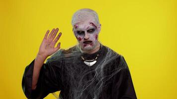 Creepy scary man with bloody scars face, Halloween zombie make-up. Scary wounded undead guy waves hand palm in hello gesture welcomes someone with hospitable expression expresses positive emotions video