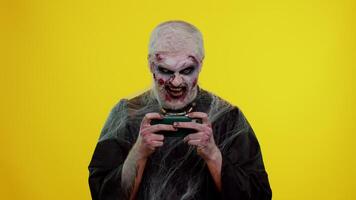Worried funny sinister man with horrible scary Halloween zombie makeup using gadget app with drive simulator game on mobile phone. Dead guy with wounded bloody scars face isolated on yellow background video