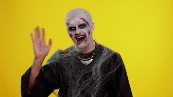 Creepy man with bloody scars face, Halloween zombie make-up. Scary wounded undead guy smiling friendly at camera and waving hands gesturing hello or goodbye, welcoming with hospitable expression video