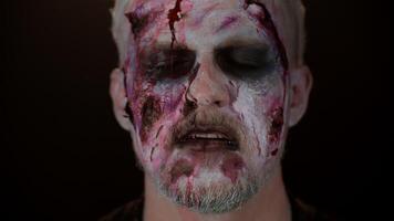 Close-up of sinister man with horrible scary Halloween zombie makeup making faces, looking ominous at camera, trying to scare, blood flows and drips on face. Dead guy with wounded bloody scars face video