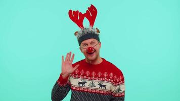 Man in Christmas sweater, deer antlers waves hand palm in hello gesture welcomes someone with hospitable expression expresses positive emotions on blue studio background. Happy New Year celebration video