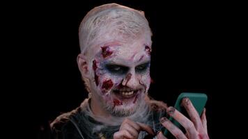 Frightening man with Halloween zombie bloody wounded makeup using mobile phone typing new post on web, sms message, browsing, addiction of social networks. Sinister undead guy on black wall background video
