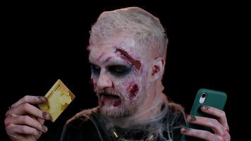 Sinister man with horrible scary Halloween zombie make-up using credit bank card and mobile phone, transferring money, purchases online shopping. Dead guy with wounded bloody scars face, black room video