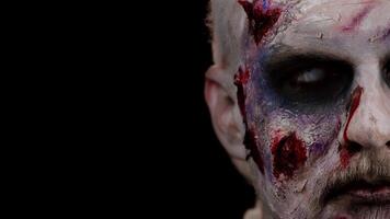 Close-up face of sinister man with horrible scary Halloween zombie makeup making faces, looking ominous at camera, trying to scare. Dead guy with wounded bloody scars face isolated on black background video
