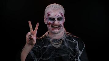 Frightening man with Halloween zombie bloody wounded makeup showing victory sign, hoping for success and win, doing peace gesture, smiling with kind optimistic expression on studio black background video