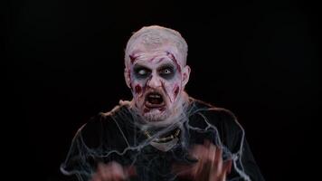 Sinister man with horrible scary Halloween zombie makeup making playful silly facial expressions and grimacing, fooling around showing tongue. Dead guy with wounded bloody scars face. Black background video