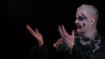 Creepy man with bloody scars face, Halloween stylish zombie make-up. Scary wounded undead guy showing thumbs up and pointing at left on blank space. Place for your advertisement logo. Sinister monster video