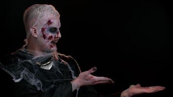 Creepy man with bloody scars face, Halloween stylish zombie make-up. Scary wounded undead guy showing thumbs up and pointing at right on blank space. Place for advertisement logo. Sinister monster video