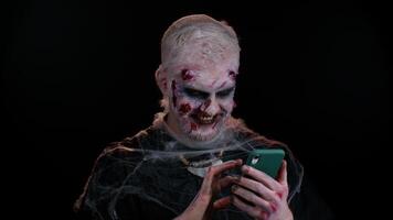 Frightening man with Halloween zombie bloody wounded makeup using mobile phone typing new post on web, sms message, browsing, addiction of social networks. Sinister undead guy on black wall background video