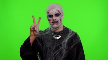 Frightening man with Halloween zombie bloody wounded makeup showing victory sign, hoping for success and win, doing peace gesture, smiling with kind optimistic expression on studio chroma key video