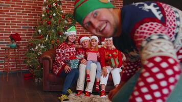 Laughing multigenerational family gathering on sofa near Christmas tree at home room waving hands hello, hi, greeting while taking selfie on smartphone on Xmas party. Distance online call video