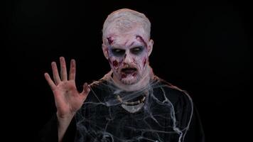 Creepy scary man with bloody scars face, Halloween zombie make-up. Scary wounded undead guy waves hand palm in hello gesture welcomes someone with hospitable expression expresses positive emotions video